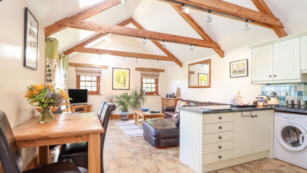 A spacious living area with a high ceiling, wooden beams, a dining table, chairs, a sofa, a kitchen with a washing machine, and a large window. - Valley View (Helston)