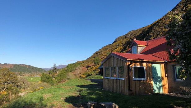 Una cabaña de madera con techo rojo, rodeada de un paisaje verde exuberante y colinas al fondo.