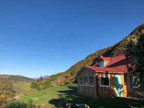 Una cabaña de madera con techo rojo, rodeada de un paisaje verde exuberante y colinas al fondo.