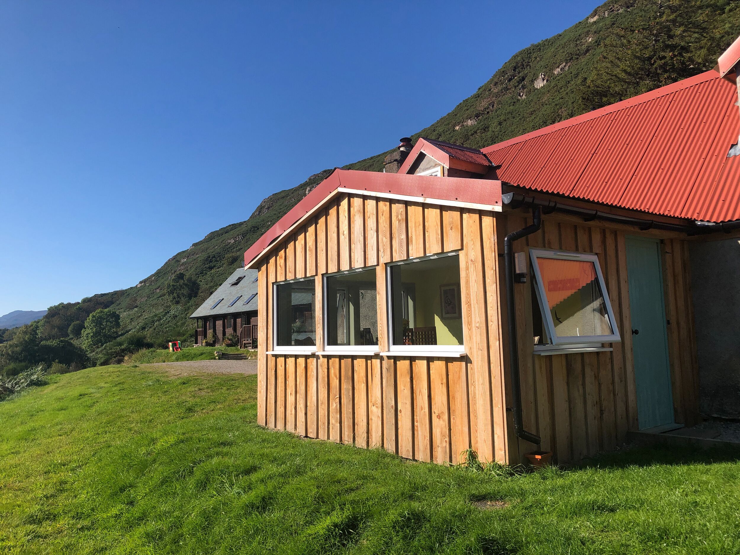 Eine Holzhütte mit rotem Dach, großen Fenstern und einer Tür, eingebettet in eine grüne Wiese mit einem Berg im Hintergrund.