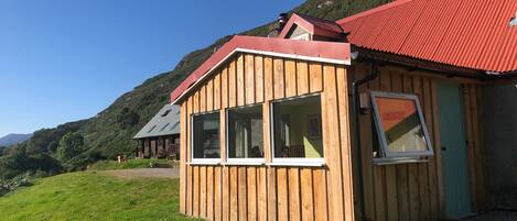 A wooden cabin with a red roof, large windows, and a door, set in a grassy area with a mountain in the background.