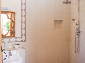 A bathroom with a hexagonal tile pattern, a shower with a handheld showerhead, a sink with a mirror above it, and a small window with a wooden frame. - High Grove (Ludlow)