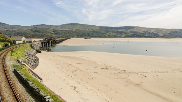 Vía de tren costera con un puente, una playa de arena y colinas al fondo.