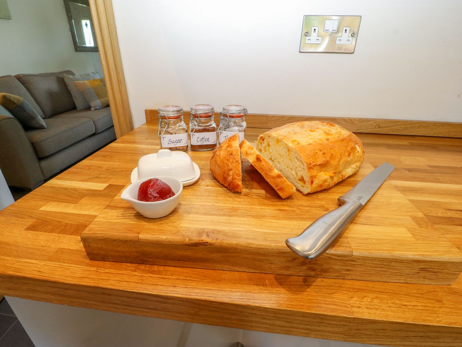 Een houten aanrecht met een brood, een mes, een schaal jam en drie gelabelde potten met suiker, koffie en thee.