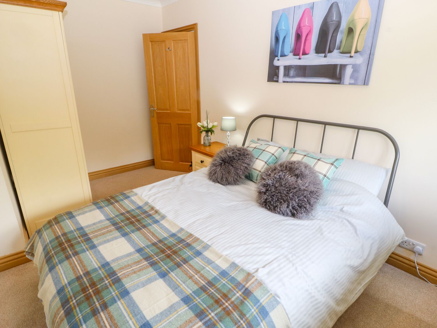 A bedroom with a bed, a wooden wardrobe, a nightstand with a lamp, and a painting of high heels on the wall.