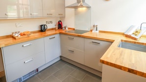 A modern kitchen with light-colored cabinets, a stainless steel range hood, a wooden countertop, and a wall-mounted clock. - Mallard Cottage (Tadcaster)