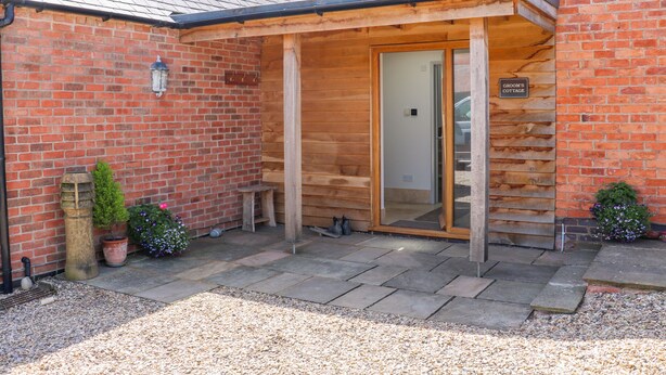 A brick cottage with a wooden porch, a door labeled "Groom's Cottage," and a small garden area with gravel and potted plants. - Grooms Cottage (Leicester)