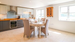 A modern kitchen with a dining area, featuring a central island, built-in oven, and ample natural light from large windows. - Grooms Cottage (Leicester)
