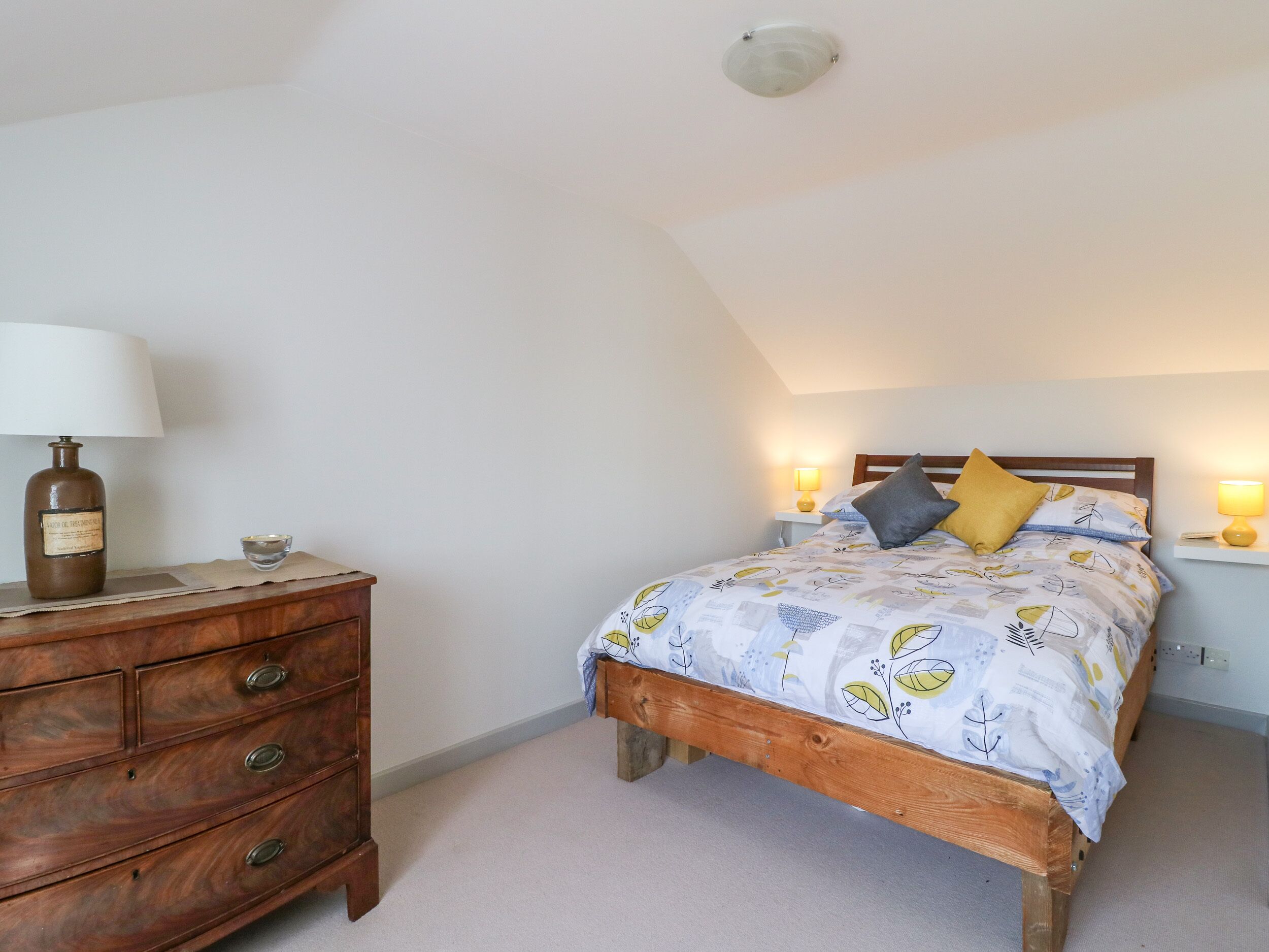 A bedroom with a wooden bed, a nightstand with a lamp, a dresser, and a ceiling light.