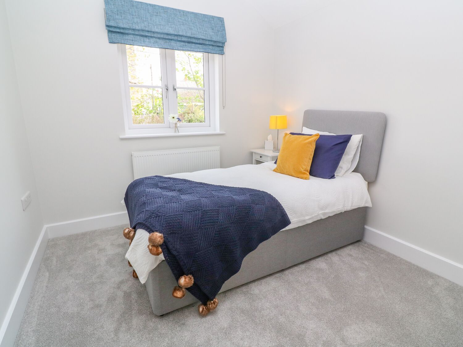 A bedroom with a bed, a bedside table, a lamp, a window with a blue valance, and a radiator.