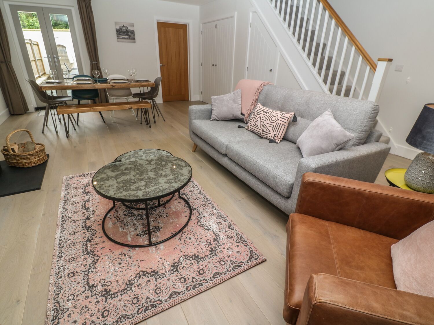 Een woonkamer met een grijze bank, een bruine leren fauteuil, een ronde salontafel en een kamerbreed tapijt met een patroon. Er staat een eettafel met stoelen in en op de achtergrond een houten deur.