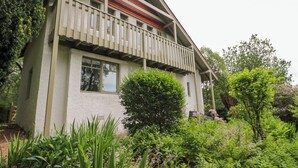 Une maison à deux étages dotée d’un balcon, entourée d’une végétation luxuriante et de plantes en fleurs.