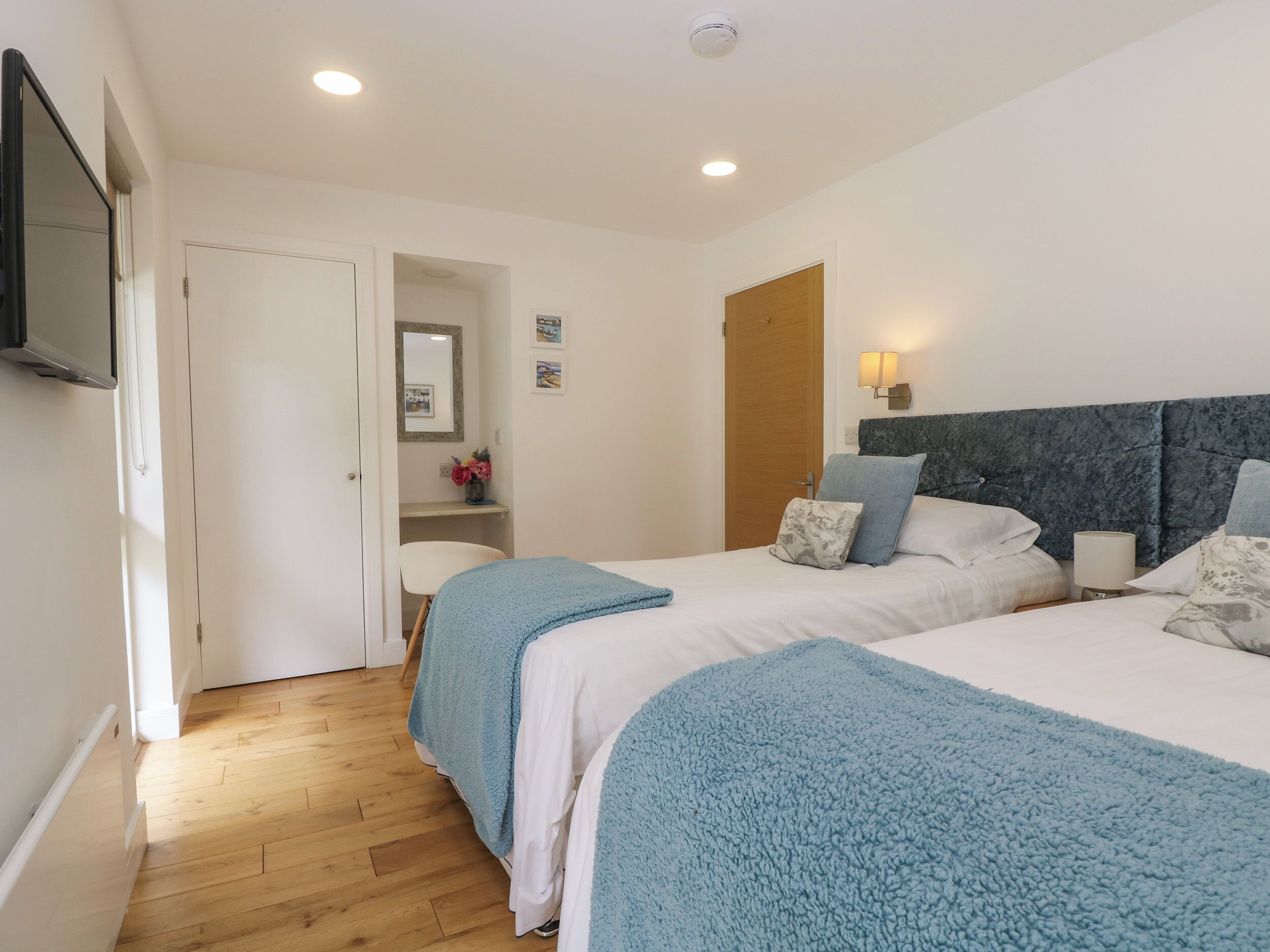 A hotel room with two beds, a wooden floor, a white door, a small white chair, a mirror, and a wall-mounted TV.
