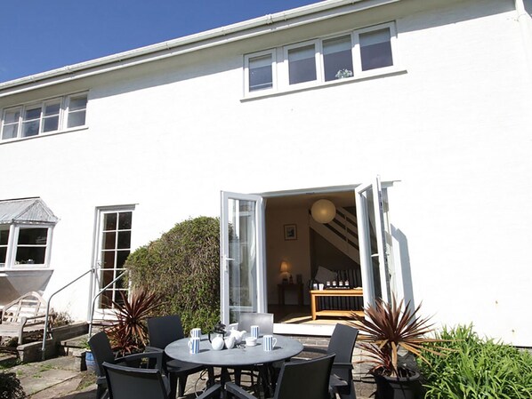 A white building with a patio area featuring a round table and chairs, a small garden, and a staircase leading to an upper level. - Bwthyn y Mor (Porthmadog)