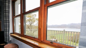 A room with a large window overlooking a lake and mountains. There are two wooden stools and a small table by the window.