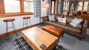 A cozy living room with a brown leather sofa, two wooden stools, a wooden coffee table, plaid curtains, and a dining area in the background.