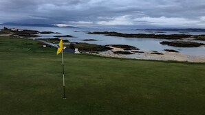 A golf course with a flagstick in the foreground, a sandy beach area, and a body of water in the background.