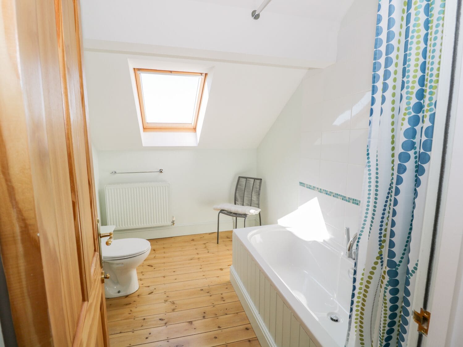 A bathroom with a wooden floor, a white bathtub, a toilet, a radiator, and a window with a wooden frame.