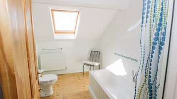 A bathroom with a wooden floor, a white bathtub, a toilet, a radiator, and a window with a wooden frame.