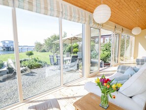 A modern room with large sliding glass doors, a wooden ceiling, and a view of a garden and water body. - Villa Marina (Barrow-in-Furness)