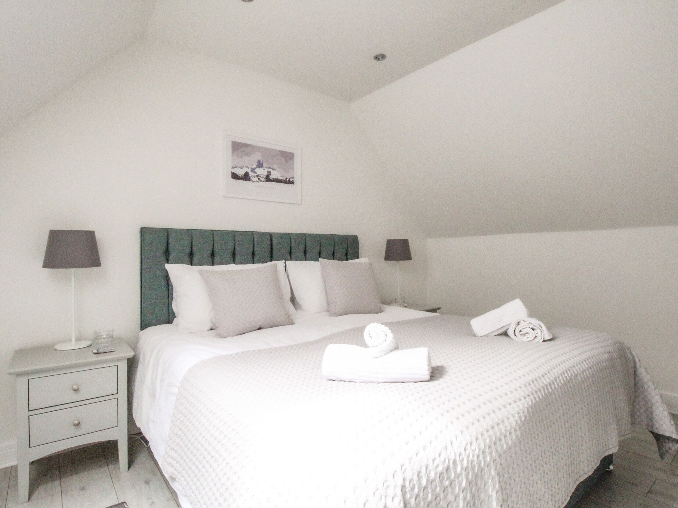 A neatly made bed with a green headboard, two bedside lamps, and a small bedside table with a lamp and a glass.