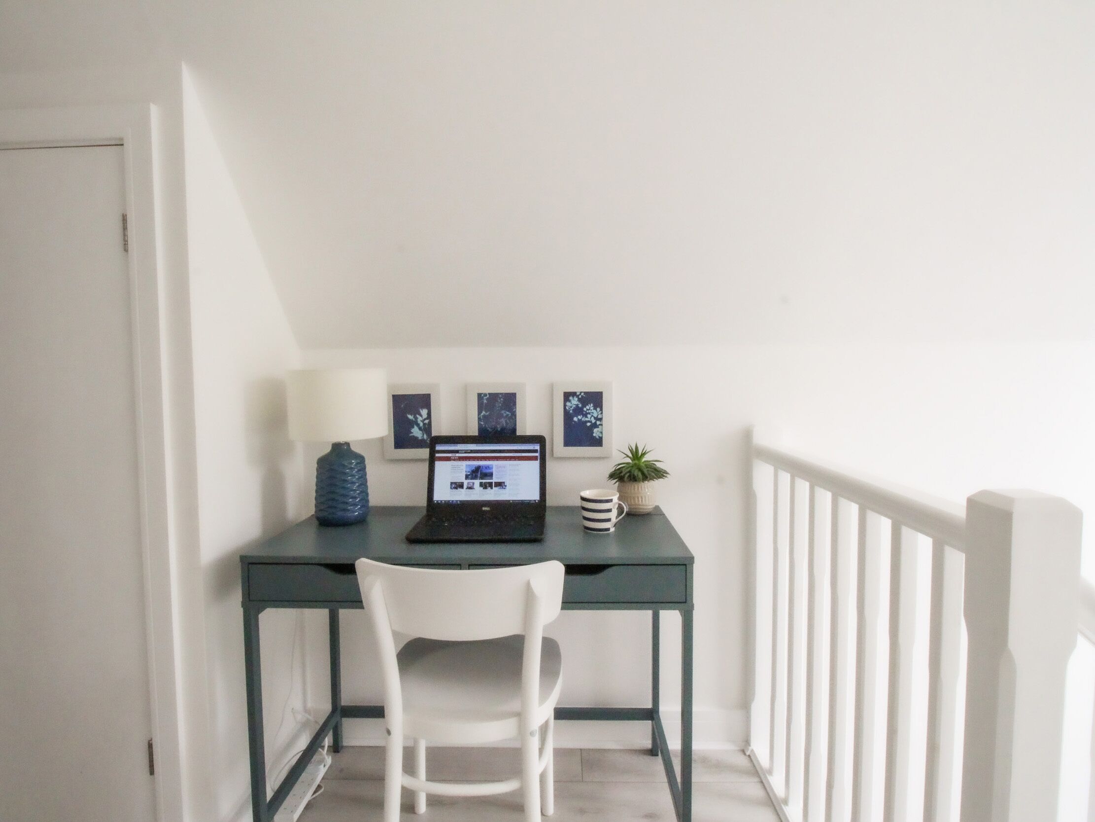 A small, white-framed desk with a laptop, a white chair, a blue vase, and a potted plant.