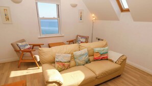 A cozy living room with a beige sofa, wooden armchairs, and a small wooden table. There are three cushions on the sofa, and a floor lamp is visible. - Meridian (St Ives)