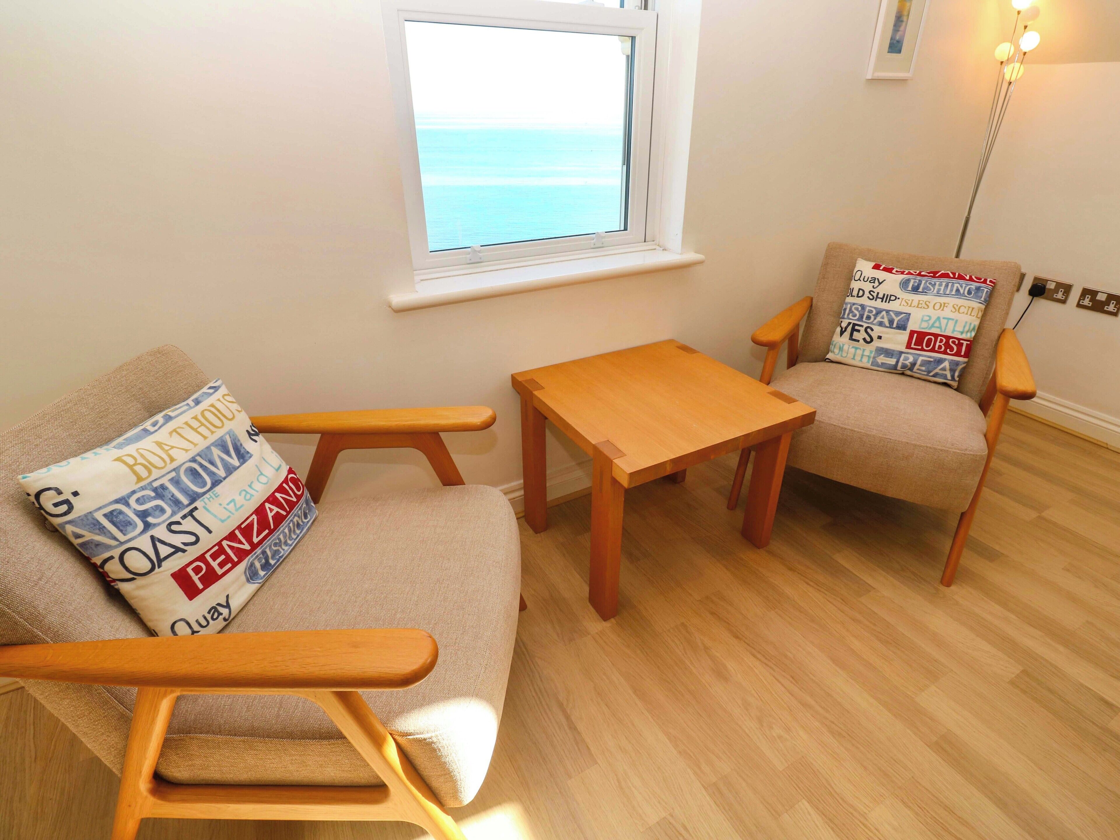 A room with a sofa, a wooden table, and a chair with a view of the sea through a window.