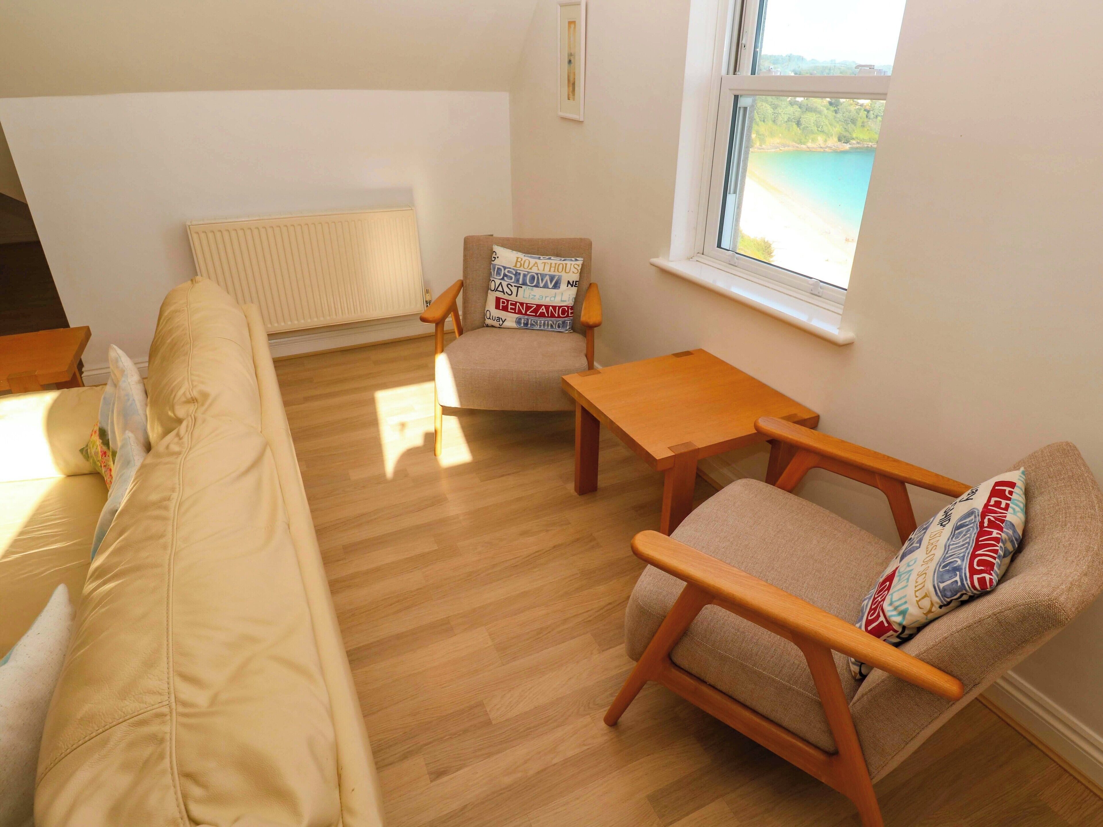 A cozy living room with a beige sofa, a wooden armchair, and a small wooden table. A window offers a view of a beach.