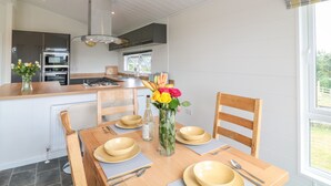 A modern kitchen with a central island, built-in appliances, and a dining area with wooden chairs and a table set for two. - Alder Lodge (Newton Abbot)