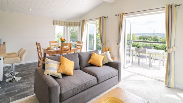 A modern living room with a grey sofa, wooden dining table, and chairs. There is a balcony with a view of greenery and a distant power line. - Alder Lodge (Newton Abbot)
