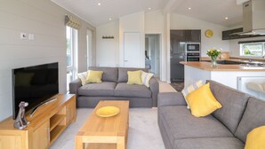 A modern living room with a flat-screen TV, grey sofas with yellow cushions, a wooden coffee table, and a kitchen area with a stove and sink. - Alder Lodge (Newton Abbot)