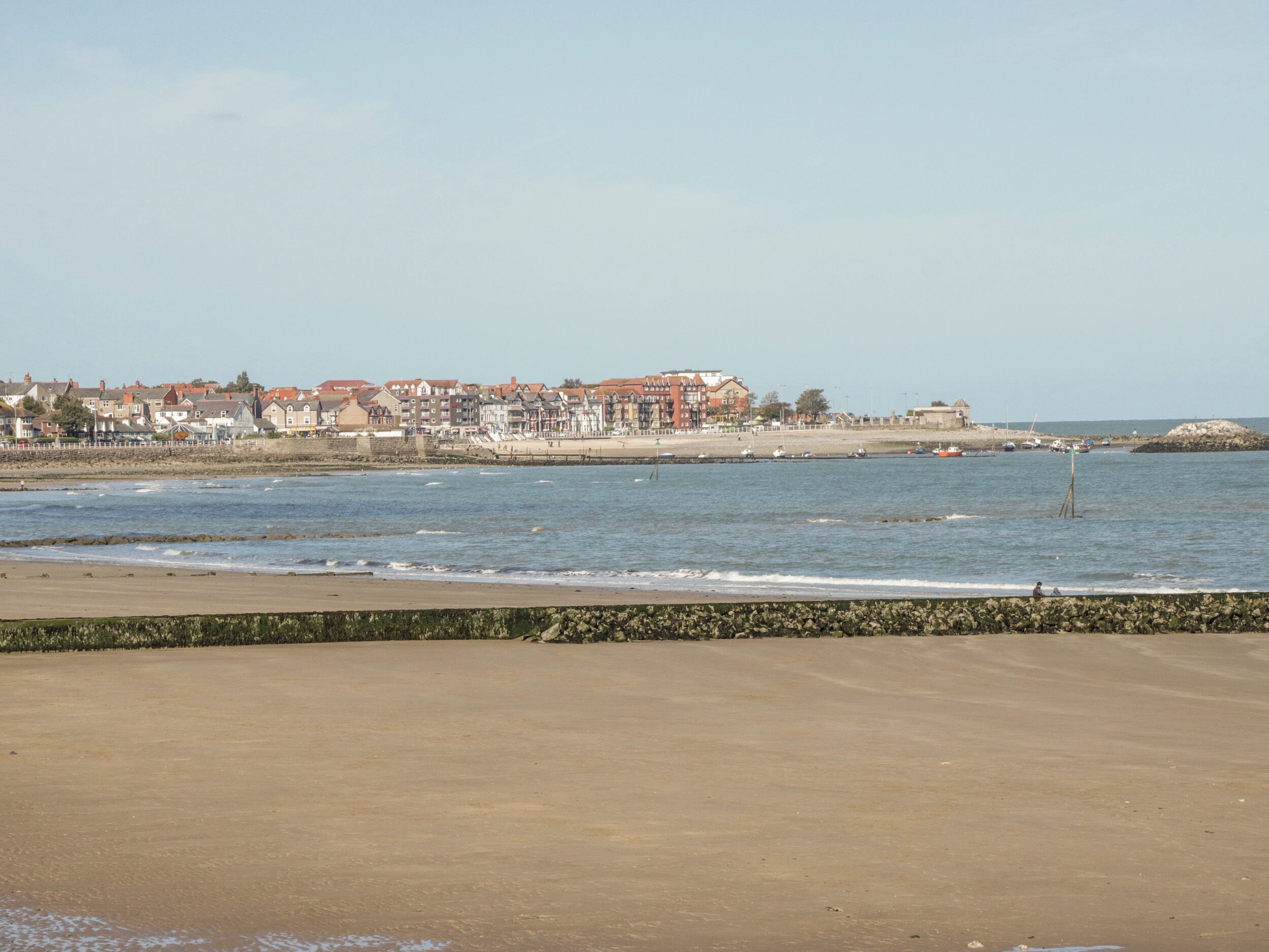 A coastal town with a sandy beach, a rocky breakwater, and buildings along the shore.