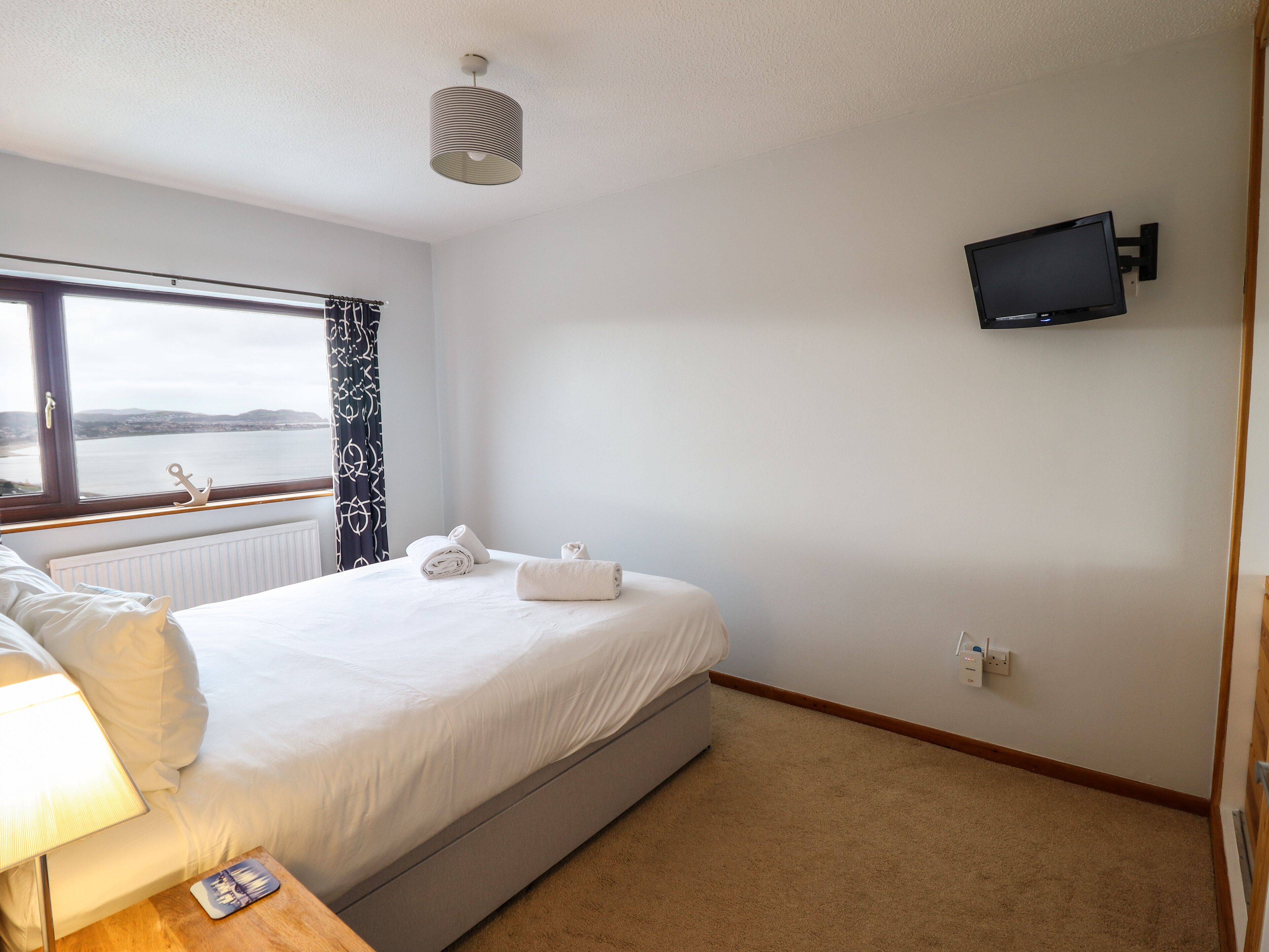 Habitación de hotel con cama, televisión en la pared, ventana con cortinas y vista al mar.
