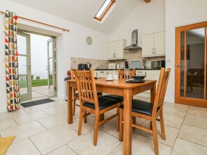 A modern kitchen with a dining table and chairs, a clock on the wall, and a view of the outdoors through a glass door. - An Gwel (Penzance)