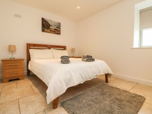 A neatly made bed with a white duvet, two bedside lamps, a wooden nightstand, a gray rug, and a painting of a coastal landscape on the wall. - An Gwel (Penzance)