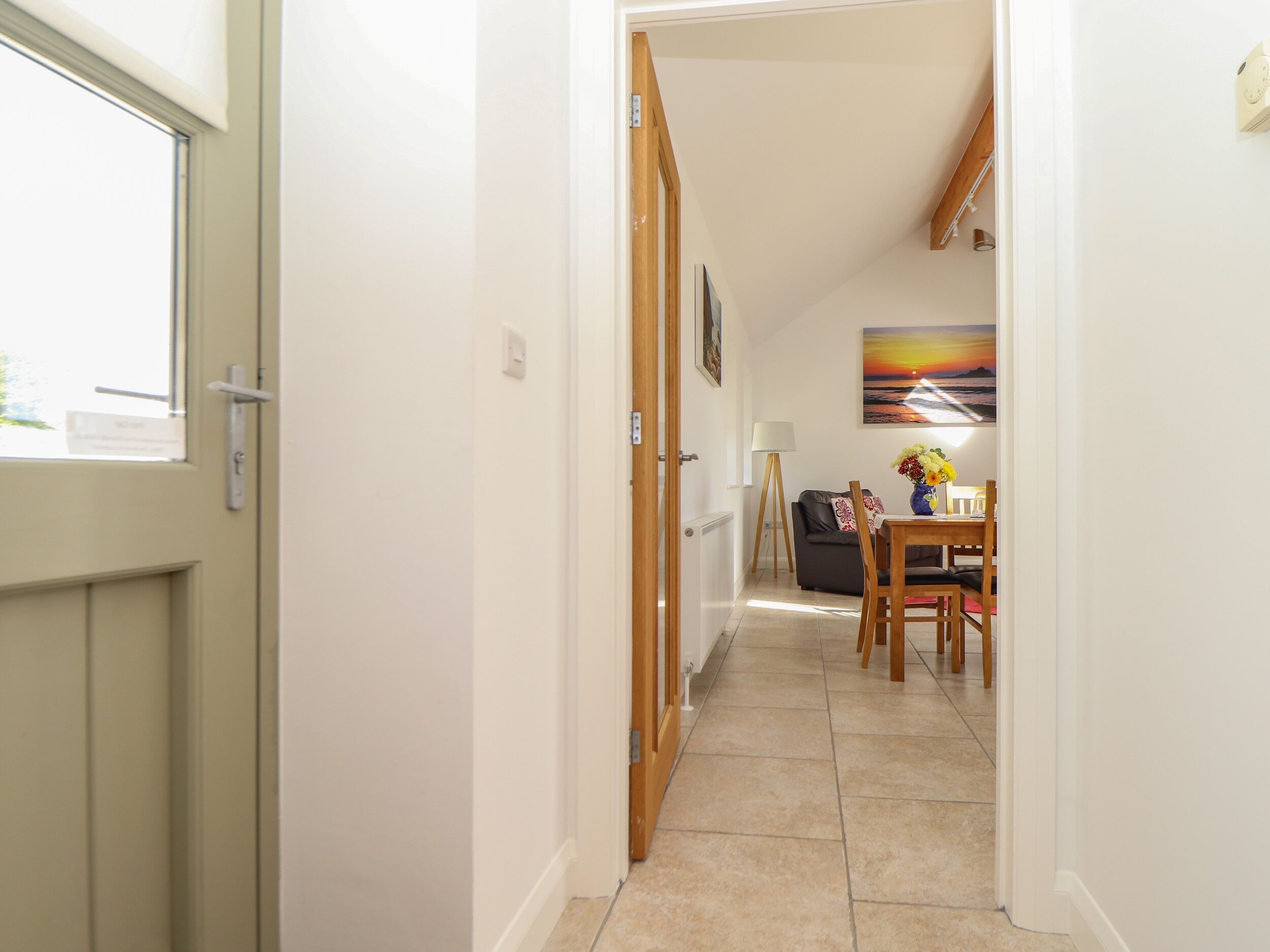 A hallway leading to a dining area with a table and chairs, a sofa, and a painting on the wall.
