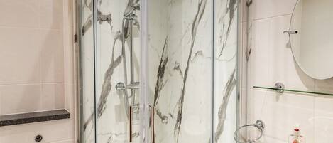 A bathroom with a glass-enclosed shower, a marble-tiled wall, a white sink, and a glass shelf.