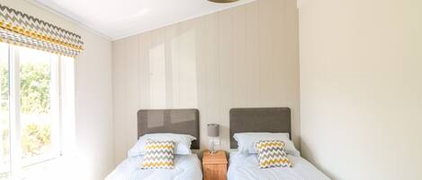A bedroom with two single beds, a wooden nightstand, a wall-mounted lamp, and a window with a patterned curtain.