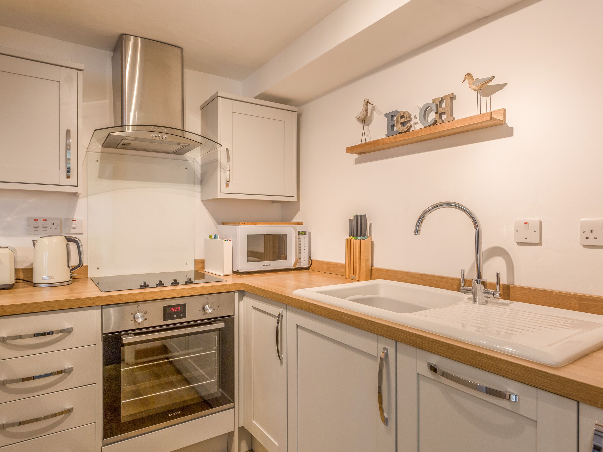 A modern kitchen with white cabinetry, a stainless steel range hood, a built-in microwave, a double sink, and a wooden shelf with decorative items.