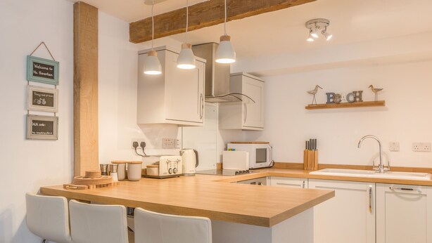 Una cocina moderna con una isla de madera, armarios blancos y taburetes de bar. Hay luces colgantes, un microondas y una cafetera en la encimera.