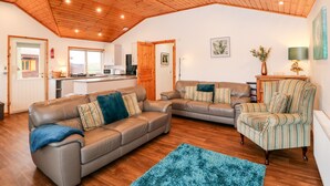 A spacious living area with a wooden ceiling, a kitchen in the background, and a striped armchair. - Teal Lodge (Lincoln)