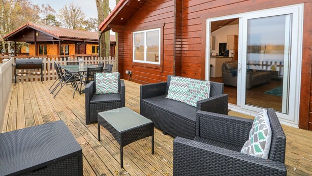 A wooden cabin with a deck, furnished with wicker furniture and a grill. - Teal Lodge (Lincoln)