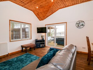 A cozy living room with a wooden ceiling, a large window with blinds, a flat-screen TV, a wooden bench, a sofa with cushions, and a sliding glass door leading to an outdoor area. - Teal Lodge (Lincoln)