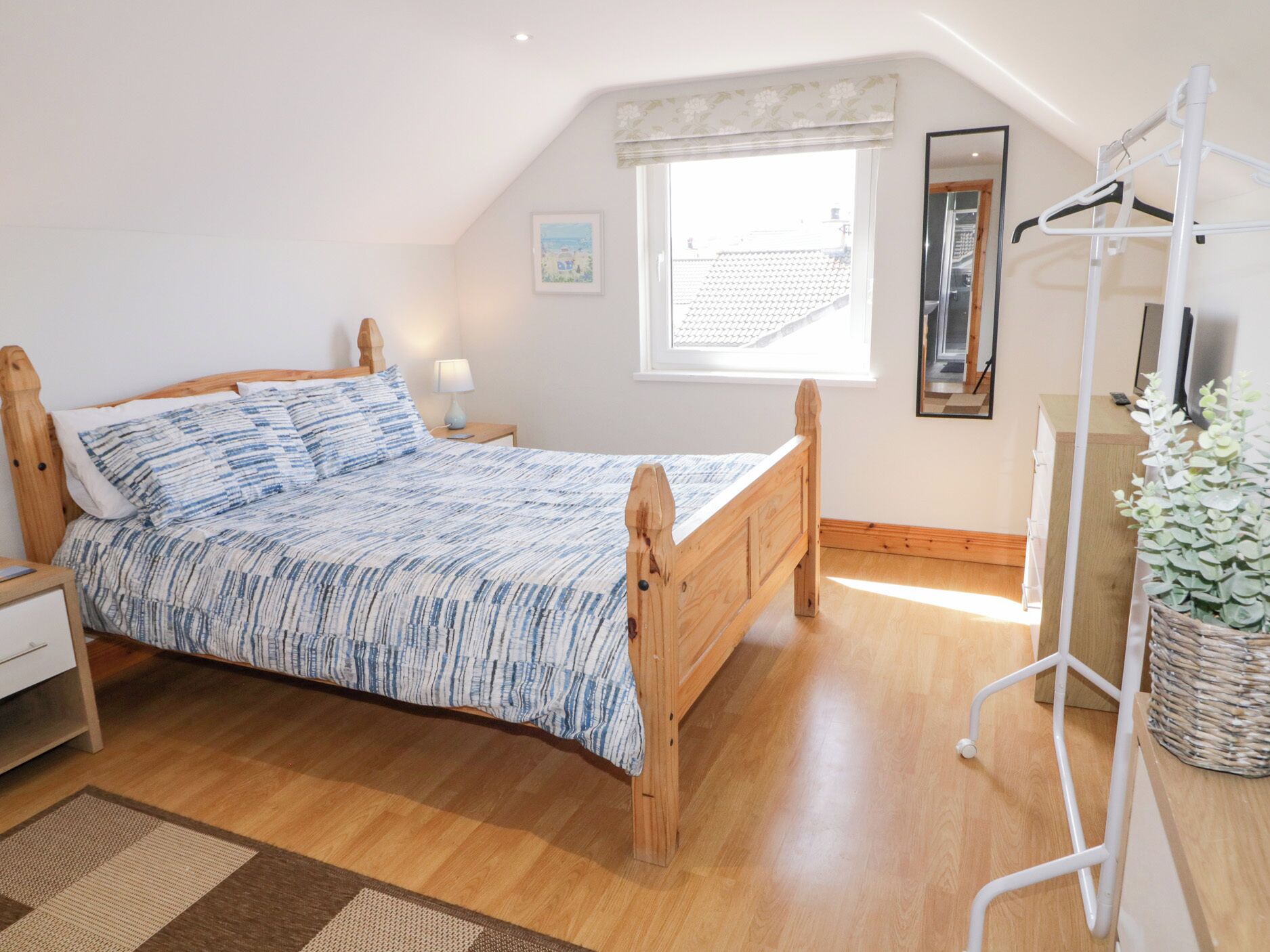 A bedroom with a wooden bed, a nightstand, a bedside table, a window with a view, a mirror, and a plant.
