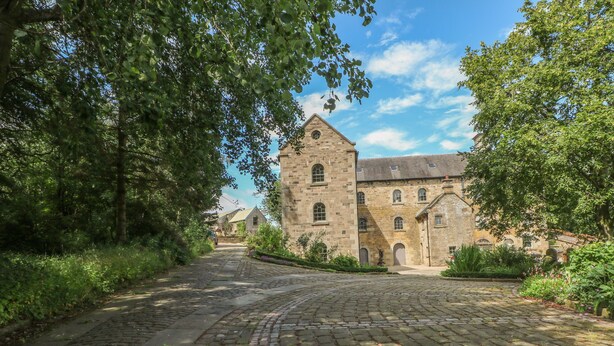 Un bâtiment historique en pierre, doté d’une tour centrale, entouré d’une végétation luxuriante et bordé d’un sentier pavé.