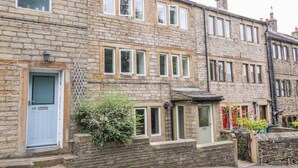 A two-story stone building with multiple windows, a visible door, and a small garden area with plants. - Bamforth Cottage (Holmfirth)