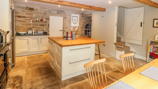 A kitchen with white cabinets, a wooden countertop, and a stone wall. There is a sink, a stove, and a chair. - Bamforth Cottage (Holmfirth)