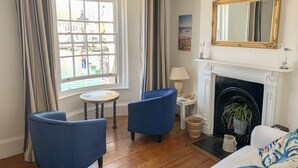 Ein Zimmer mit Kamin, zwei blaue Sessel, ein weißes Sofa und ein großes Fenster mit Vorhängen.