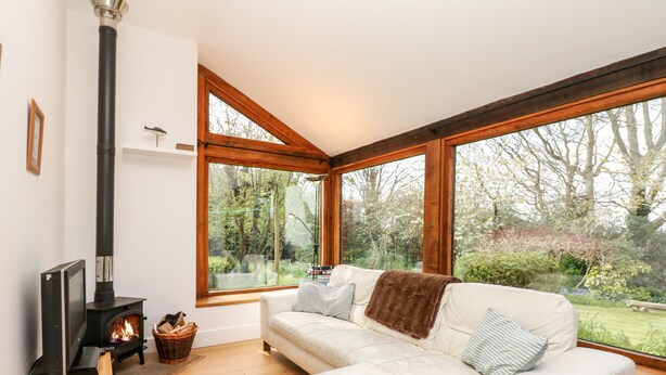 A cozy living room with a white sofa, a fireplace, and large windows overlooking a garden. - Puffin Cottage (Axminster)